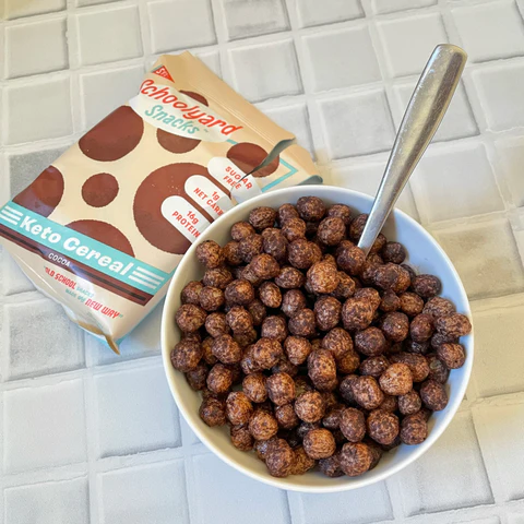A bowl of Schoolyard Snacks Chocolate Keto Cereal with a spoon beside its packaging on a tiled surface.