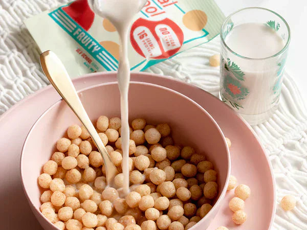 Milk being poured into a bowl of Schoolyard Snacks fruity cereal with a packet and a glass of milk nearby.