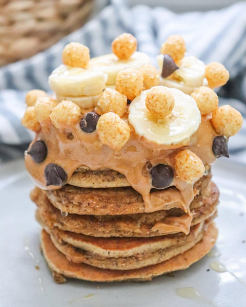 Stacked pancakes topped with peanut butter, banana slices, and Schoolyard Snacks protein cereal puffs.