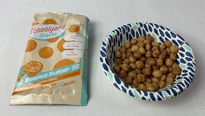 **Alt text:** A packet of Schoolyard Snacks Peanut Butter Protein Cereal next to a bowl filled with the round cereal pieces on a patterned blue and white dish.