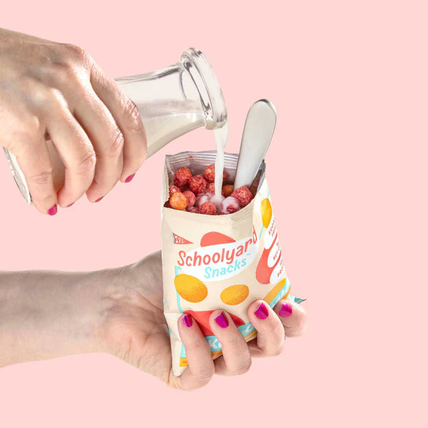A hand holding a Schoolyard Snacks cereal pouch filled with red cereal balls, while milk is being poured into it from a glass bottle, with a spoon inside.